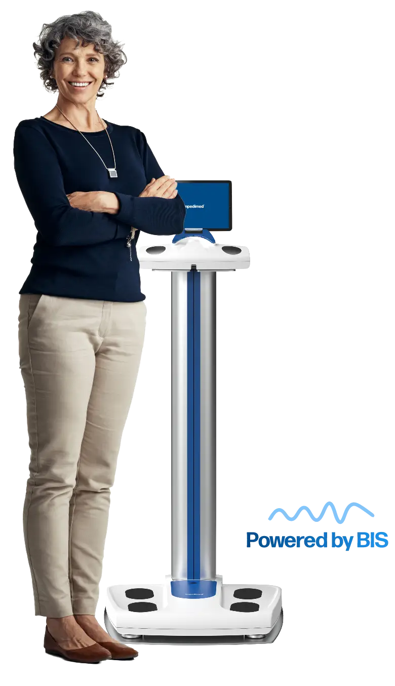 Smiling woman in a dark blue top and beige pants stands with arms crossed beside a tall medical scale with a screen, logoed device labeled “Powered by” in the lower right.